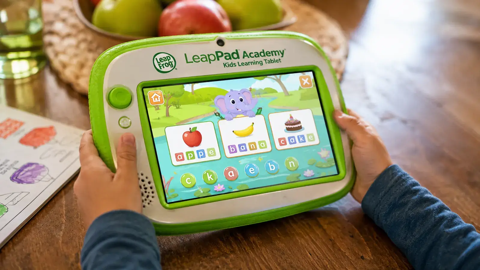 Child's hands holding the LeapFrog LeapPad Academy tablet showing a colorful learning app on screen